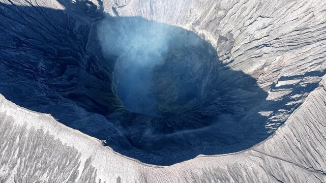 Deep blue crater dominates aerial view. Smoke rises gently from volcanic lake center at Bromo