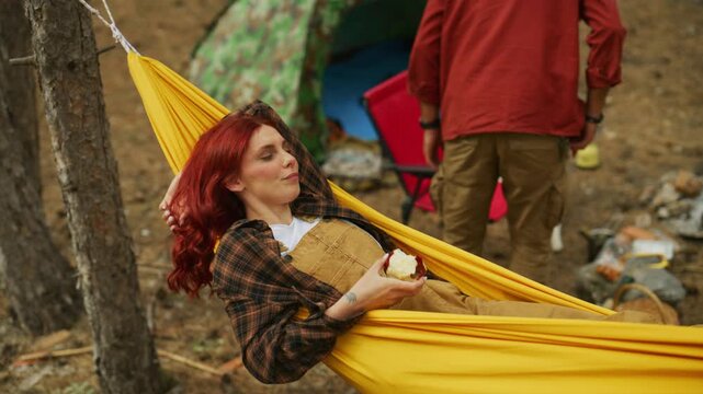 Woman holds an apple and smiles in a yellow hammock while a man moves near the tent at a forest campsite. Handheld footage captures camping leisure.