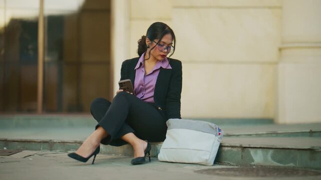 A young Asian businesswoman intently writes in her notebook while using her phone outdoors. She is focused on her work or studies in an urban setting.