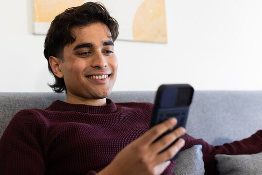 African American man reclining on gray couch at home in maroon sweater, holding phone and smiling