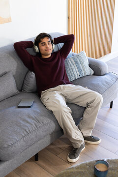African American man reclining on gray sofa at home wearing maroon sweater and over-ear headphones