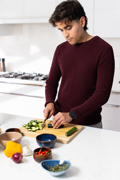 Asian man wearing maroon sweater slicing cucumber with chef's knife at white kitchen counter