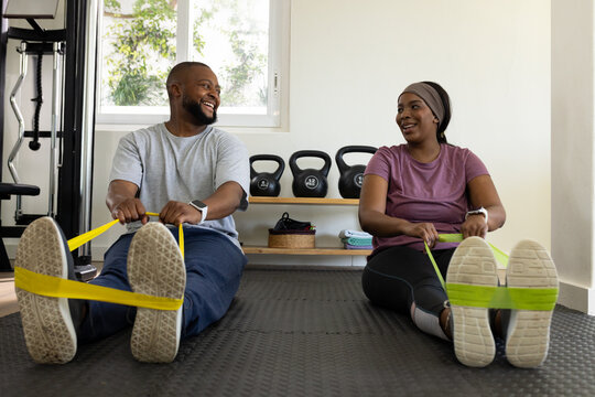 African couple exercising, rowing seated with yellow and green bands on mat at home gym