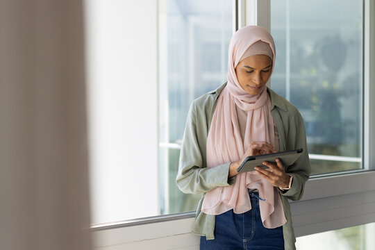 Female standing in modern building by floor-to-ceiling glass window, tapping tablet with smartwatch