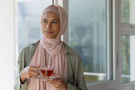 Adult African woman standing near glass window holding glass teacup and saucer in light-pink hijab