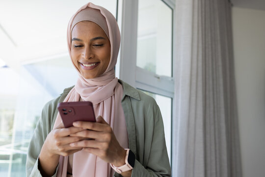 African American woman using maroon phone at home by window wearing pink hijab and smartwatch