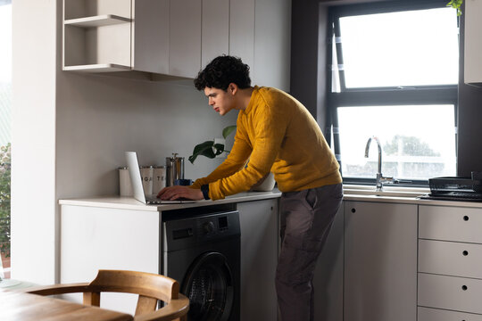 African American man leaning and typing on laptop at counter near washer in mustard sweater
