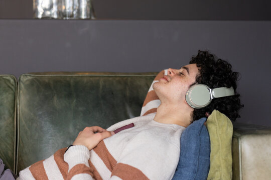 African American man lying on green couch in over-ear headphones holding phone at chest, copy space