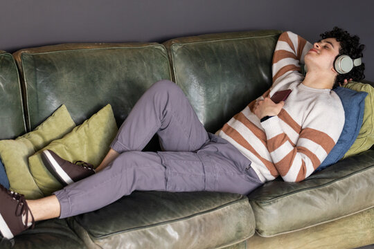 African American man reclining on green sofa, wearing sweater, holding smartphone with headphones