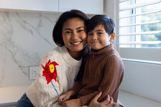 Diverse mid adult mother and child sitting on kitchen counter by window holding red pinwheel toy