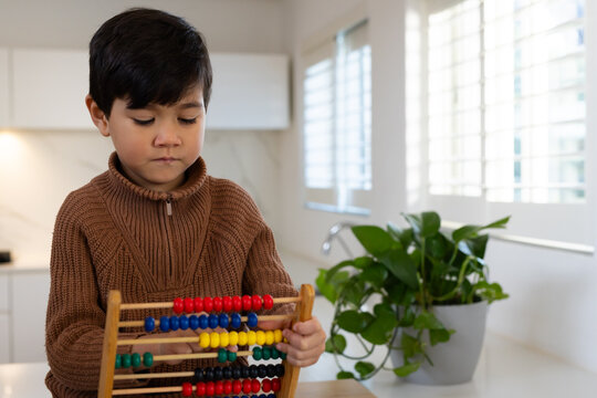Asian schoolboy in brown sweater holding wooden abacus at kitchen counter near potted plant
