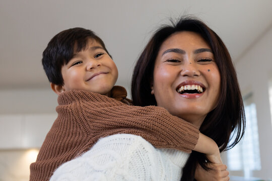 Diverse mother and youth son playing piggyback, laughing at home near blinds in knit sweaters