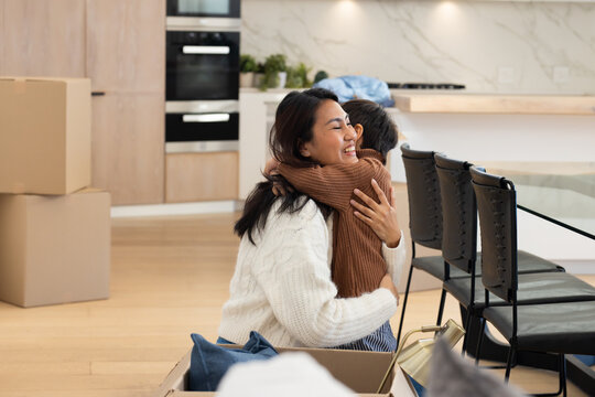 Asian mother wearing white sweater and child kneeling hugging in kitchen among moving boxes