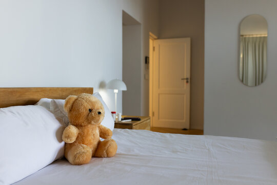Light-brown teddy bear is sitting upright on white bed in bedroom near bedside table and lamp