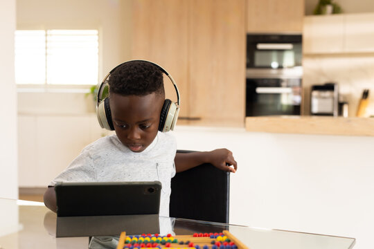 School-age male child sitting at glass table in kitchen wearing headphones using tablet with abacus