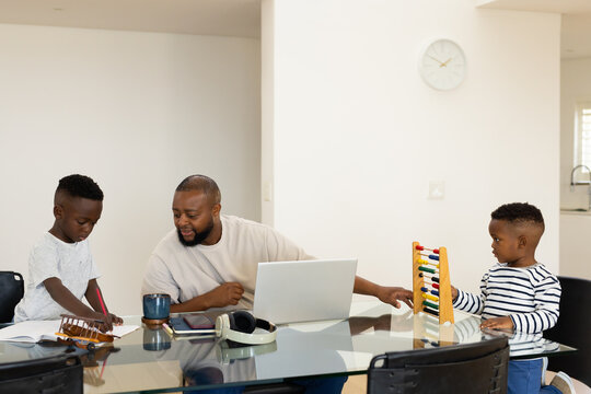African American father guiding two boys at glass dining table at home using laptop and abacus