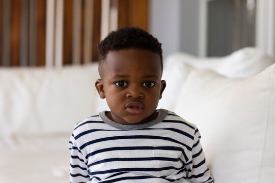 African male toddler sitting on white sofa wearing striped shirt near wooden slats and pillows
