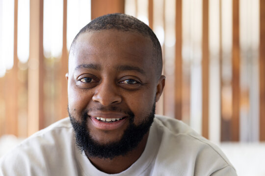 African American man smiling and sitting on sofa by wooden slats at home, wearing light sweatshirt