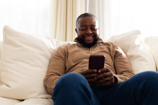 African American man reclining on sofa at home wearing tan sweater holding smartphone, showing ring