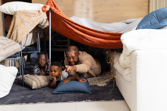 African-American dad, two sons in loungewear lying in orange throw fort at home, lights on