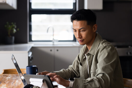 Asian man sitting at wooden table in modern kitchen using laptop and tablet, blue mug