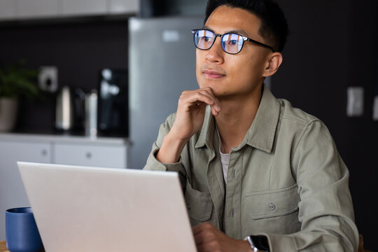 Asian man sitting at kitchen table, resting chin, using laptop with mug, wearing glasses