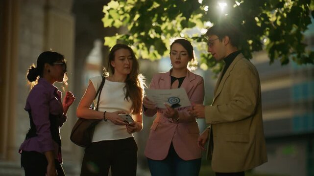 A diverse group of business professionals discusses project data outdoors under bright sunlight. Colleagues collaborate, reviewing documents and a smartphone in a natural, urban setting.