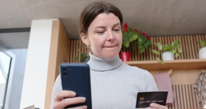 Adult woman sitting on tan couch holding smartphone and card, checking payment then picking tea