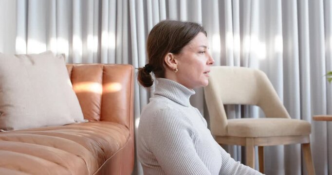 Woman sitting beside sofa rubbing temples from headache, in turtleneck with smartwatch, easing pain