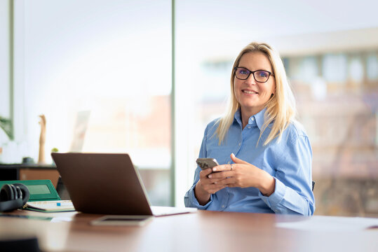 Multitasking businesswoman holding a smartphone while working on a laptop in a modern office