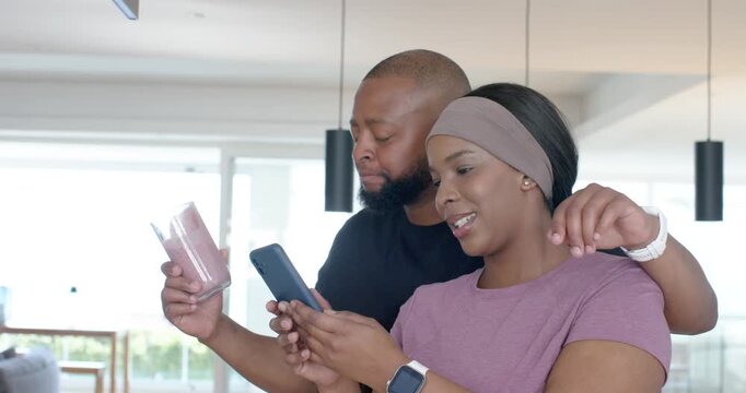 Mature couple leaning in kitchen, woman scrolling blue smartphone, drawing man holding pink tumbler