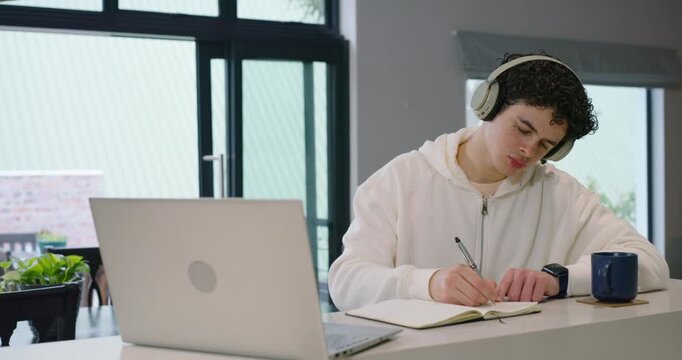 African American man sitting at counter wearing headphones, writing while checking laptop for info