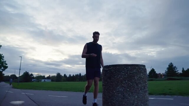 Low angle wide shot of a tired young athlete walking past a round concrete bollard after finishing a run in a public park in slow motion.