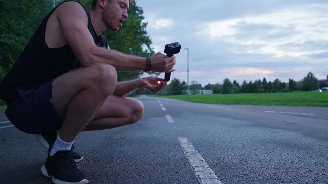 Young male runner crouches on an asphalt park route and unfolds an action camera grip into a mini tripod before filming his workout in slow motion.