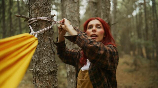 Red-haired woman secures a yellow hammock with rope on a pine tree, then walks away through the autumn forest while camping.