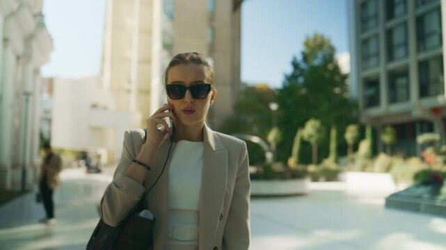 A businesswoman wearing sunglasses walks through a modern urban park while engaged in a phone call. The professional woman communicates on her smartphone in a contemporary city environment.