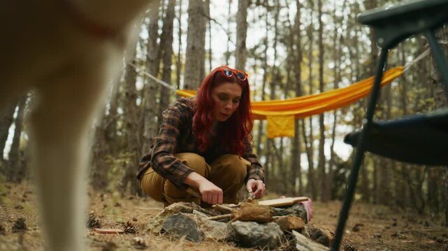 Red-haired woman builds a campfire by arranging kindling in a fire pit while camping at a pine forest campsite.