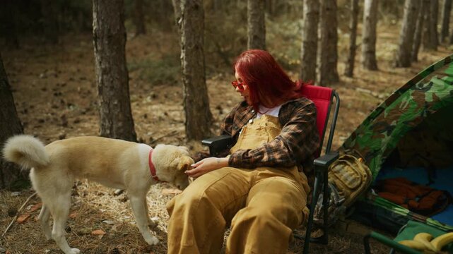 Woman pets and cuddles her dog, placing sunglasses on its head while camping beside a tent in the forest.