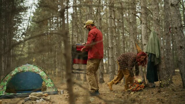 Couple sets up camp in a pine forest as the man unfolds a camping chair and woman packs gear beside a tent.