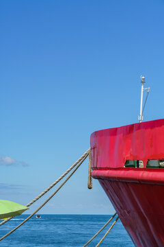 Ship bow moored with natural hemp ropes against a clear blue sky and calm sea. Close-up of a vessel prow anchored in a harbor with traditional mooring lines. Large red vessel. Maritime transportation.