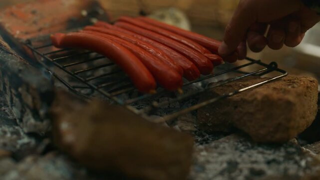 Sausages cook over glowing coals as a hand adjusts food on a campfire grill during a smoky outdoor barbecue.