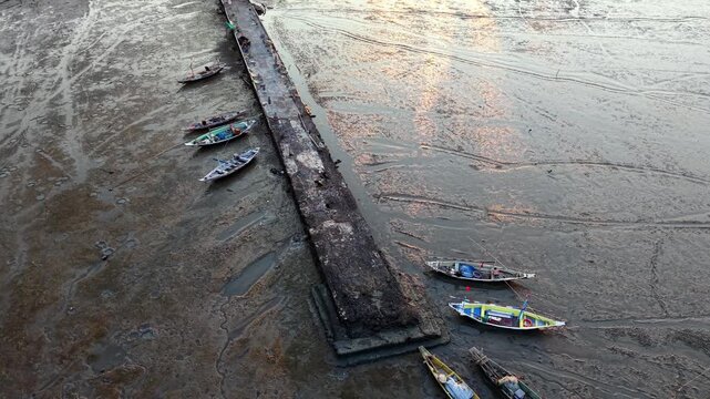 Sunlight glints on muddy river surface. Boats rest near a weathered wooden pier at Surabaya coastal