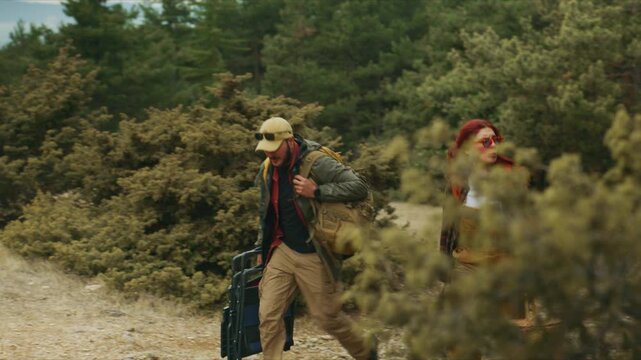 Couple hikes uphill through a pine forest carrying camping gear, a backpack, picnic basket and folding wagon toward a hilltop destination.