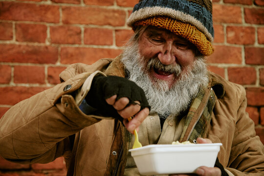 Senior Caucasian homeless man with beard smiling while eating food from container outdoors. Image suitable for charity campaigns, social welfare programs, poverty awareness materials