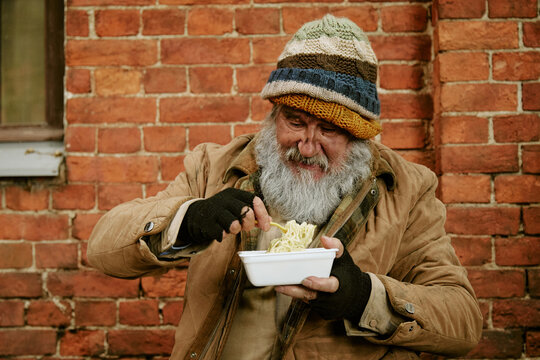 Senior Caucasian homeless man eating hot noodles outdoors near brick wall. Visual concept for charity foundation campaign, social support programs, volunteer food distribution