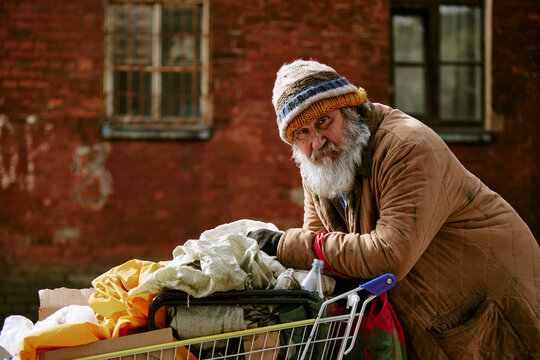 Portrait of senior Caucasian man leaning on shopping cart outdoors representing homelessness and poverty. Suitable for social charity campaigns, medium shot