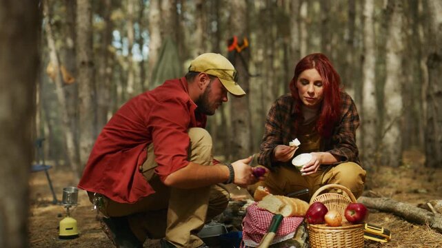 Couple eats food and prepares a camping meal beside a picnic basket in a sunny pine forest.