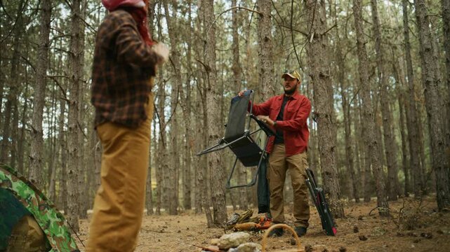 Man sets up portable camping chairs at a forest campsite while another camper prepares nearby in the woodland.