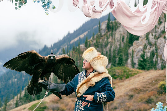 Almaty, Kazakhstan - 19 October 2026: View of a traditional Kazakh falconer in a fur hat and embroidered coat, holding a golden eagle on his gloved arm against rocky mountain slopes.