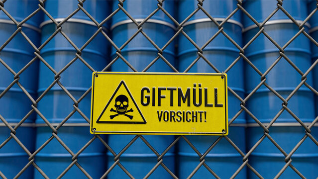 Warnschild Giftm&uuml;ll Vorsicht an Absperrzaun vor gestapelten F&auml;ssern